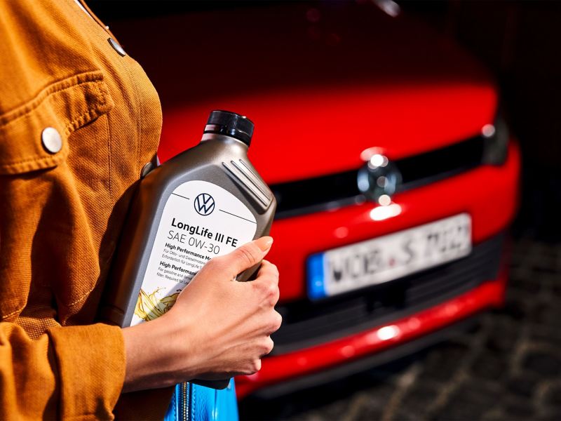 Eine Frau steht vor einem Volkswagen und hält eine Motorenölflasche in der Hand