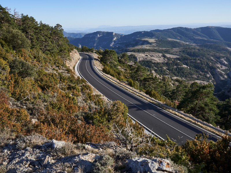 Vue panoramique d'une route sinueuse traversant un paysage montagneux, avec une végétation luxuriante verte et brune sur les pentes et un ciel clair. La route s'éloigne au loin, révélant d'autres sommets et vallées.