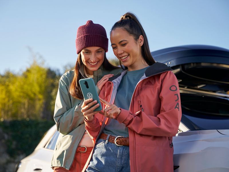 Zwei Frauen lehnen an einem VW Auto mit Dachbox und schauen sich VW Newsletter auf Smartphone an