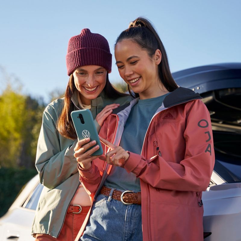 Zwei Frauen lehnen an einem VW Auto mit Dachbox und schauen sich VW Newsletter auf Smartphone an