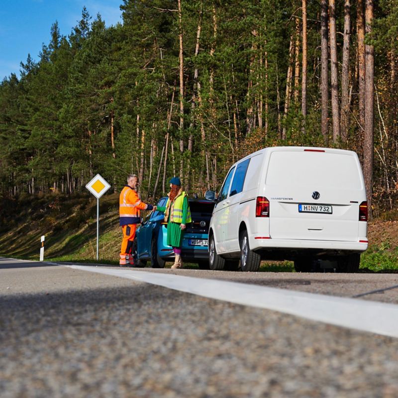 Ein VW Servicemitarbeiter von der Pannenhilfe hilft einer Frau mit ihrem liegen gebliebenen Auto, im Vordergrund ist ein Warndreieck