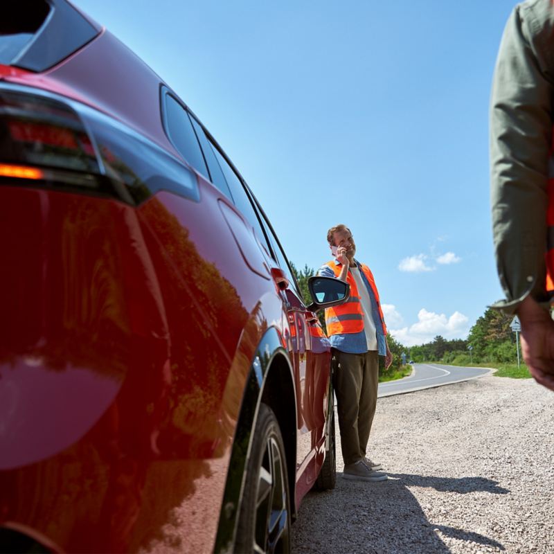 Mann in Warnweste ruft neben rotem VW Modell die VW Notdienst Nummer an