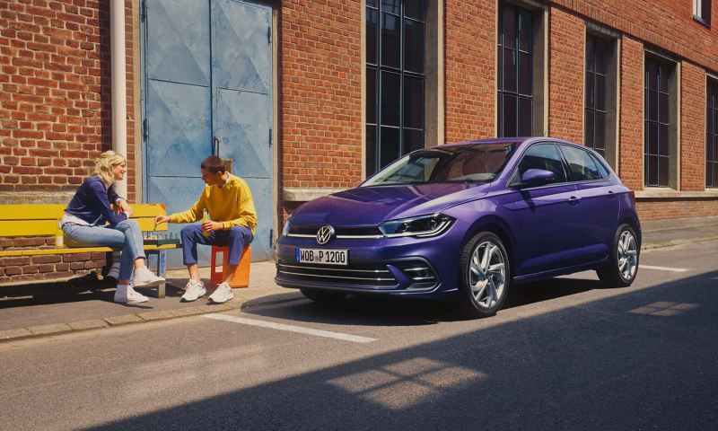 Two people are sitting on a bench, a VW Polo is parked on the side of the road