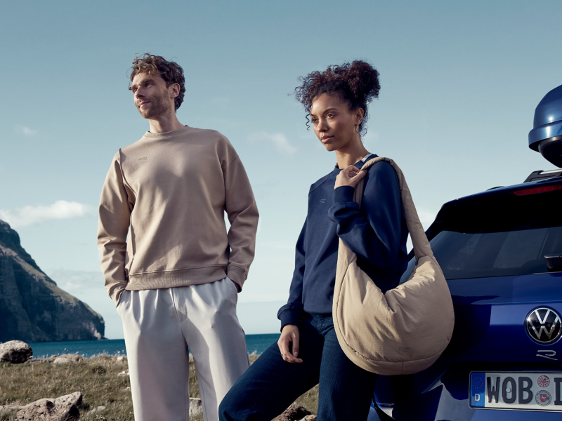 Mann und Frau mit Kleidung aus der R-Kollektion stehen neben Heck eines VW Golf R, Meer und Klippen im Hintergrund