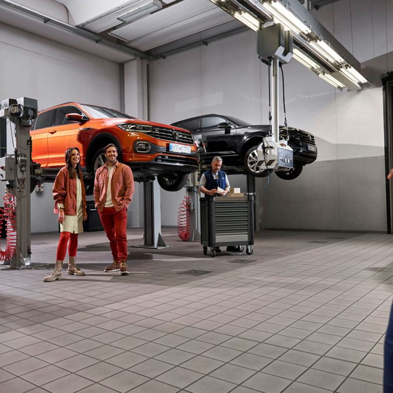 Two customers and a VW service employee in the workshop, VW T-Cross and VW Touareg on lifting platforms