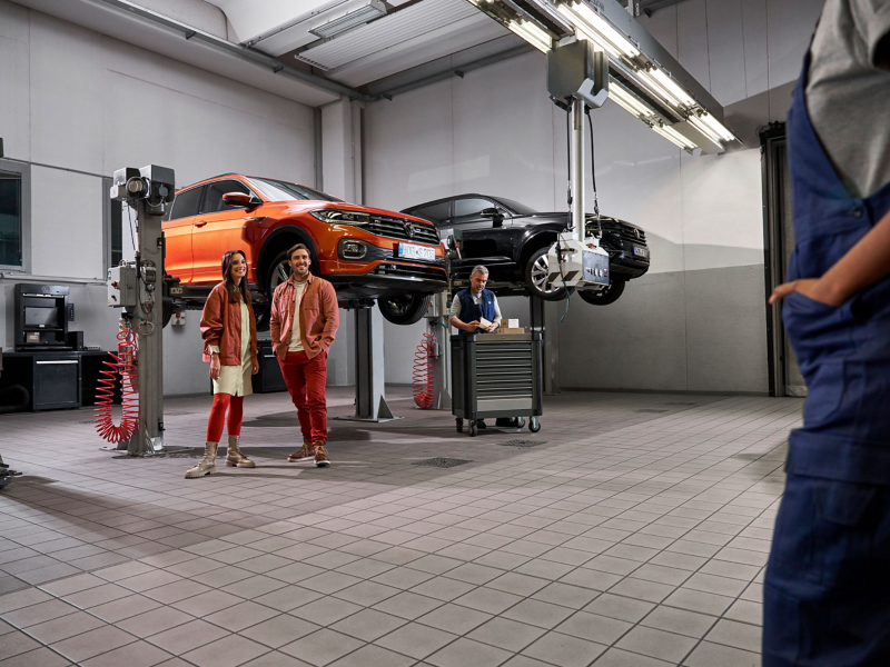 Due clienti e un addetto all’assistenza Volkswagen in officina, una Volkswagen T-Cross e una Volkswagen Touareg sulle piattaforme di sollevamento