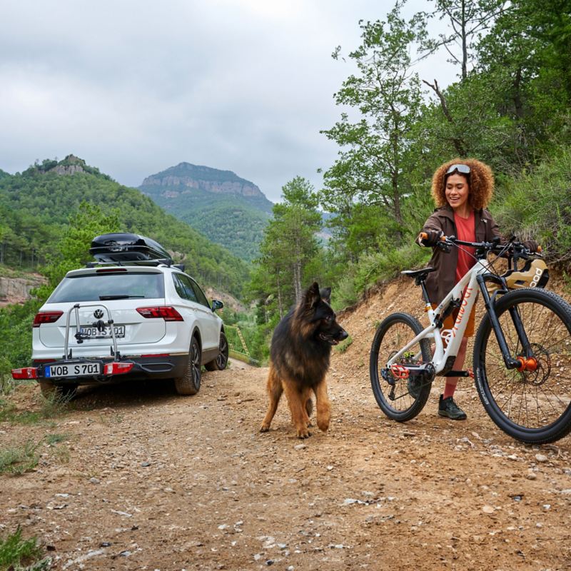 Eine Frau mit einem Fahrrad und einem Hund an ihrer Seite, im Hintergrund ein VW Auto