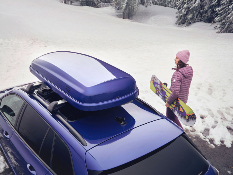 Frau mit Snowboard neben VW Auto mit Zubehör Dachbox in Winterlandschaft