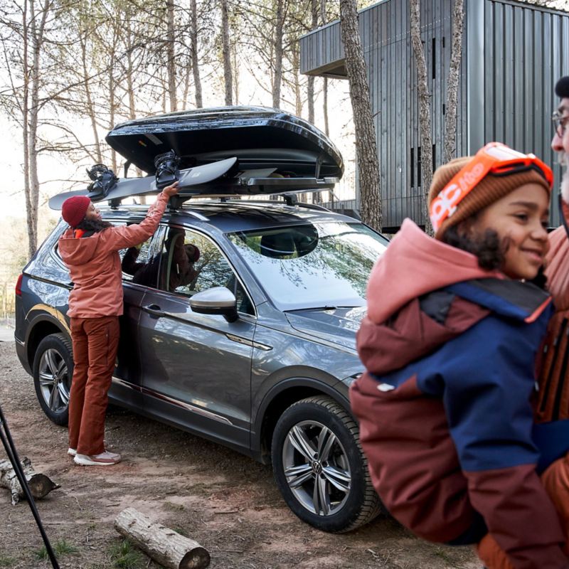 Frau verstaut Skier in VW Zubehör Dachbox, im Vordergrund Mann mit Kind auf dem Arm
