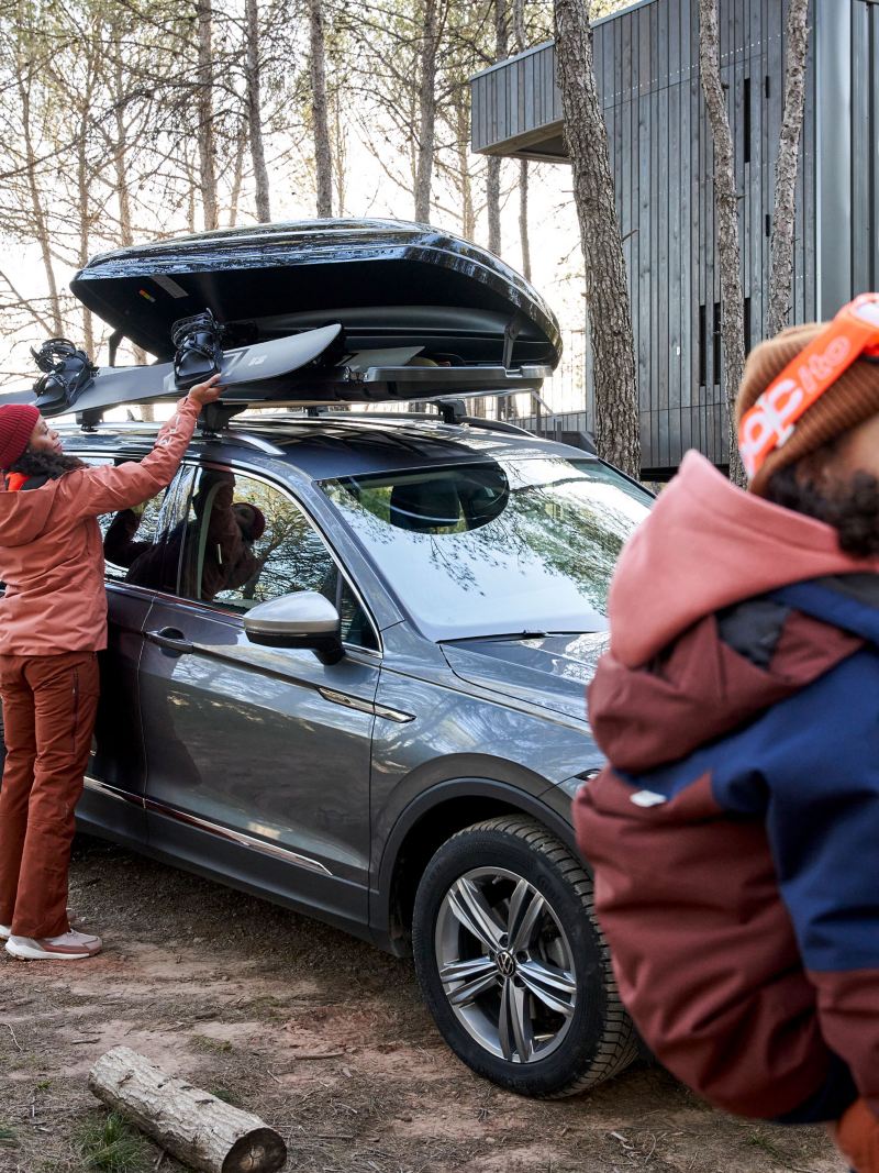 Frau verstaut Skier in VW Zubehör Dachbox, im Vordergrund Mann mit Kind auf dem Arm