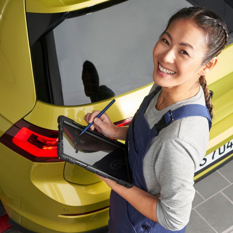 Eine glückliche VW Servicemitarbeiterin mit einem Tablet in der Hand bei der Inspektion eines Autos