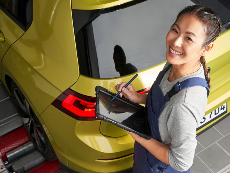 Glückliche VW Servicemitarbeiterin mit Tablet in der Hand bei der Inspektion eines Autos