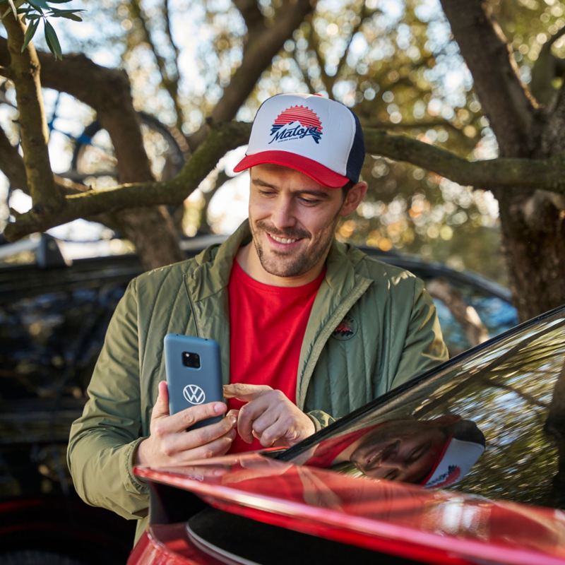 Mann steht neben einem elektrischen VW Auto und schaut auf Smartphone – VW Serviceanfrage