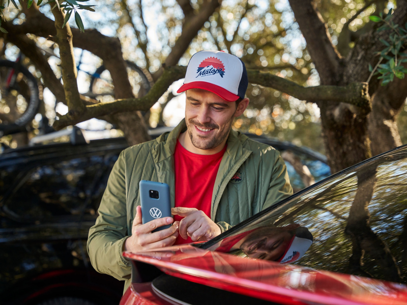 Mann steht neben einem elektrischen VW Auto und schaut auf Smartphone – VW Serviceanfrage