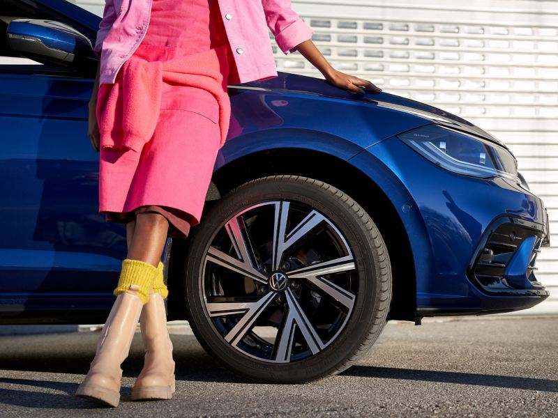 A woman in summer clothes leans against her blue VW vehicle – focus on a VW summer tyre