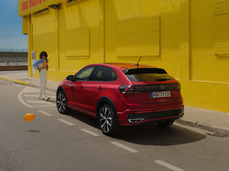 VW Taigo in Rot am Straßenrand vor Gebäude, eine Frau geht auf das Fahrzeug zu