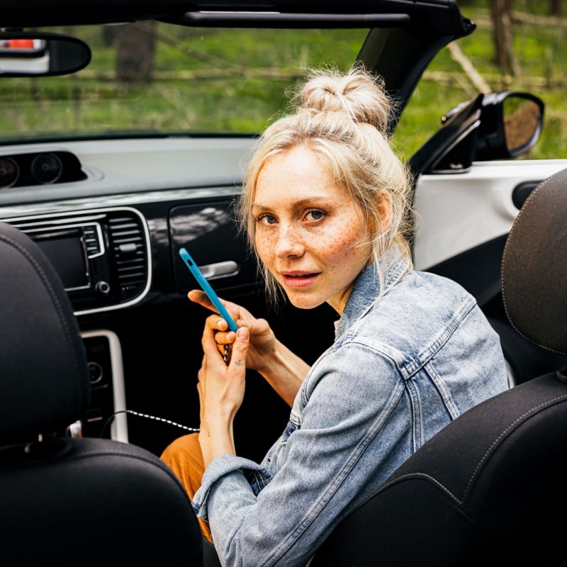 A blonde woman in the passenger seat of a VW convertible with a smartphone in her hand
