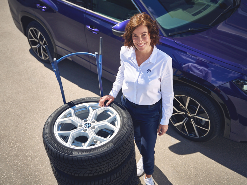 View from above of VW service employee standing next to sack truck with stacked wheels, VW Tayron in the background
