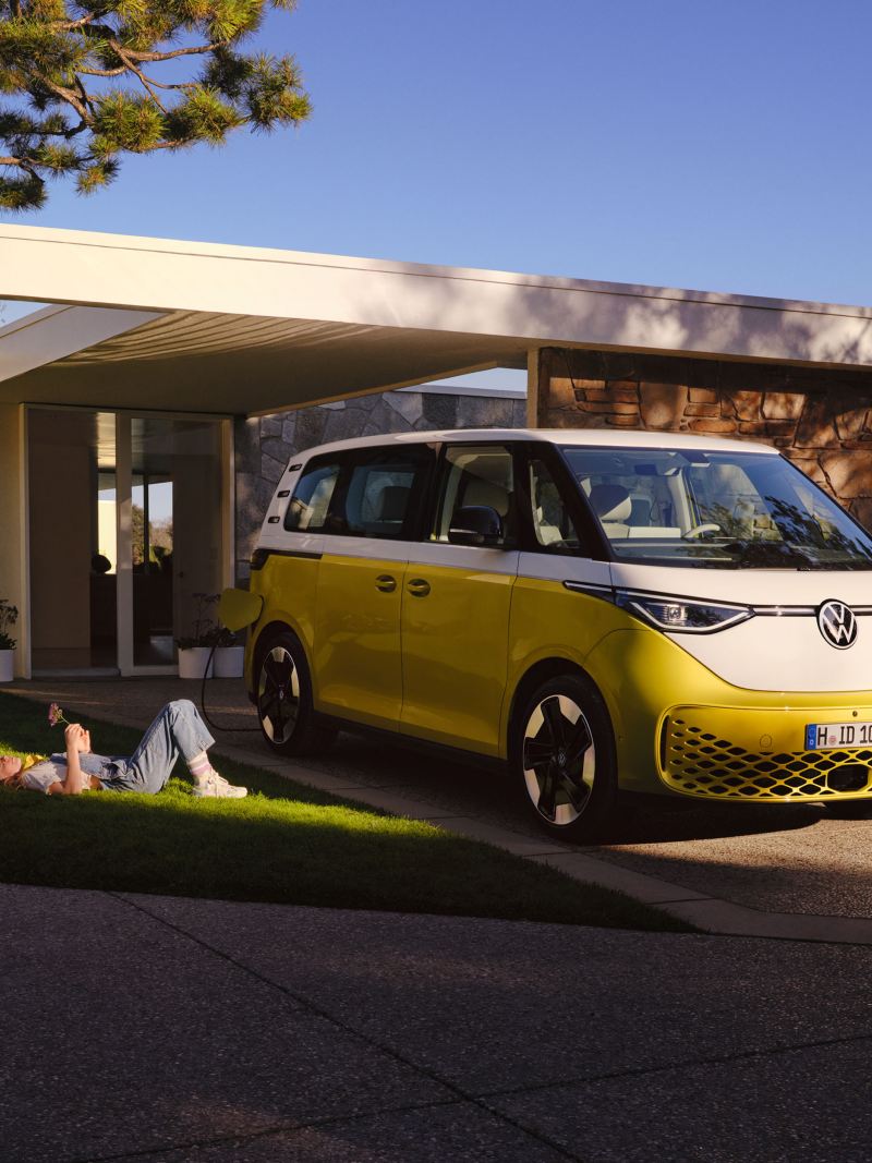 Ein weiß-gelber VW ID. Buzz steht auf einer Einfahrt, umgeben von einer Familie