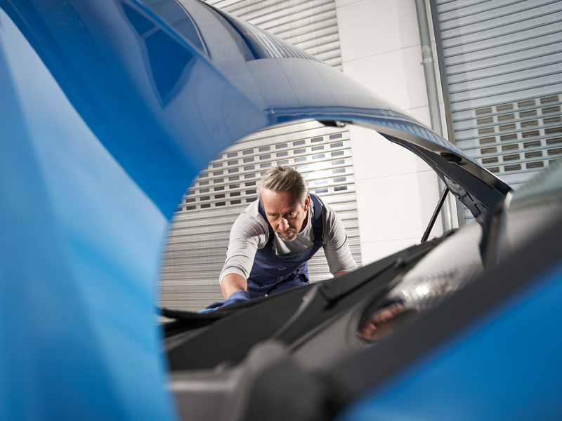 Un mécanicien avec une barbe de trois jours, portant un uniforme de travail, se penche sur le moteur ouvert d'une voiture bleue dans un garage.