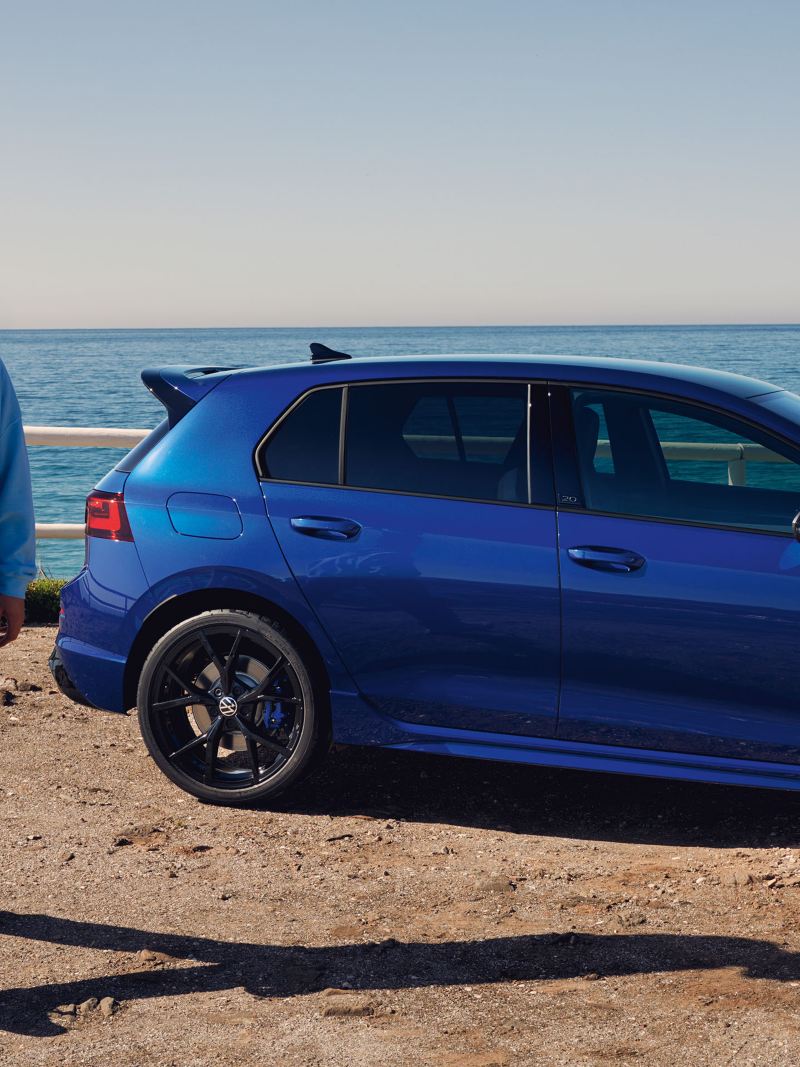 Ein Mann steht vor seinem blauen VW Golf R „20 Years“ – im Hintergrund das Meer