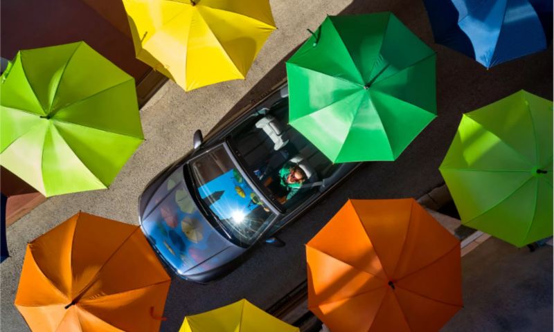Convertible VW driving among colorful umbrellas.