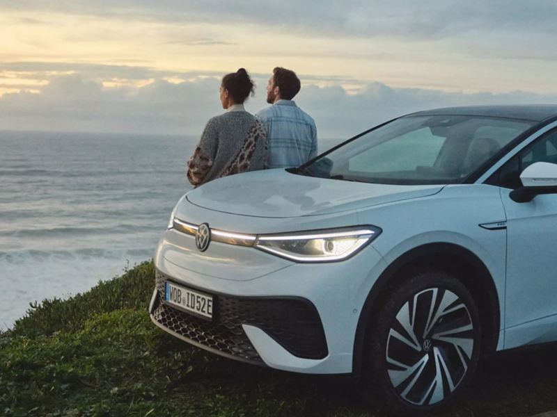 Couple stands by electric vehicle looking at ocean.