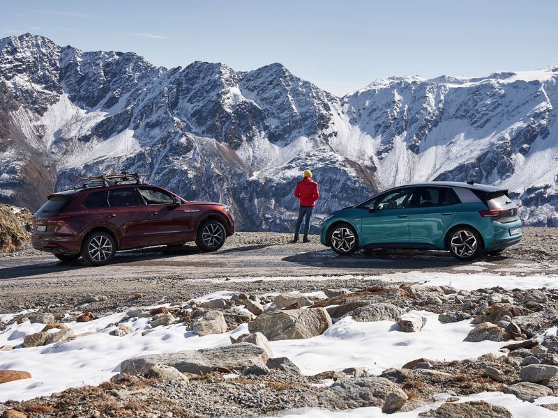 Un hombre disfruta de la vista de un paisaje montañoso cubierto de nieve, a su lado dos coches VW – neumáticos de invierno