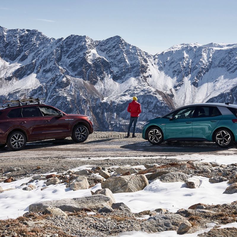 Een man geniet van het uitzicht op een besneeuwd berglandschap, naast hem twee VW’s – winterbanden
