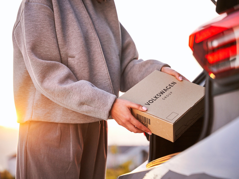 Frau verstaut Päckchen, auf dem ein Volkswagen-Schriftzug zu sehen ist, im Kofferraum eines VW Autos