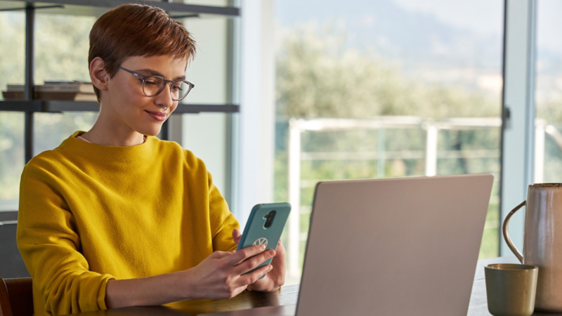 Frau schaut vor ihrem Laptop auf Ihr Handy und prüft, wo ihr Volkswagen gerade ist.