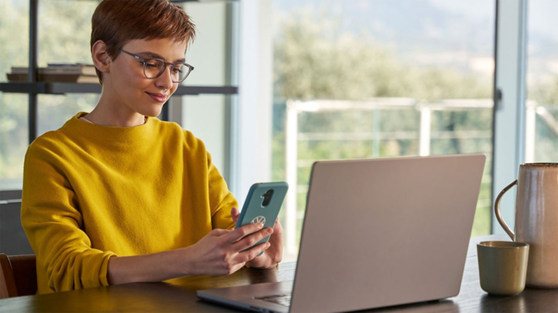 En person sitter framför en dator med en mobiltelefon i handen