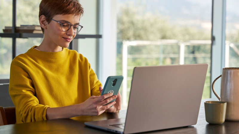 Frau sitzt an einem Tisch mit Handy in der Hand, auf dem Tisch steht ein geöffneter Laptop.