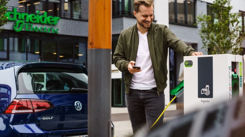 Chico usando su móvil apoyado en una estación de carga conectado a un Volkswagen e-Golf azul
