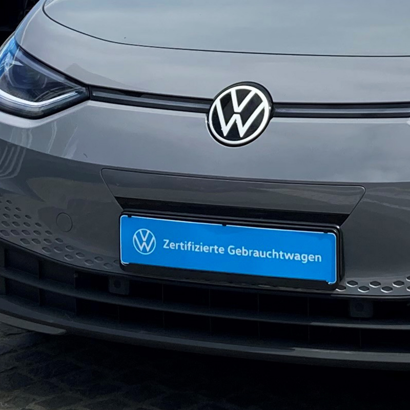 Die Vorderseite eines grauen VW mit dem Logo in Großaufnahme, darunter ein blaues Nummernschild mit der Aufschrift „Zertifizierte Gebrauchtwagen“