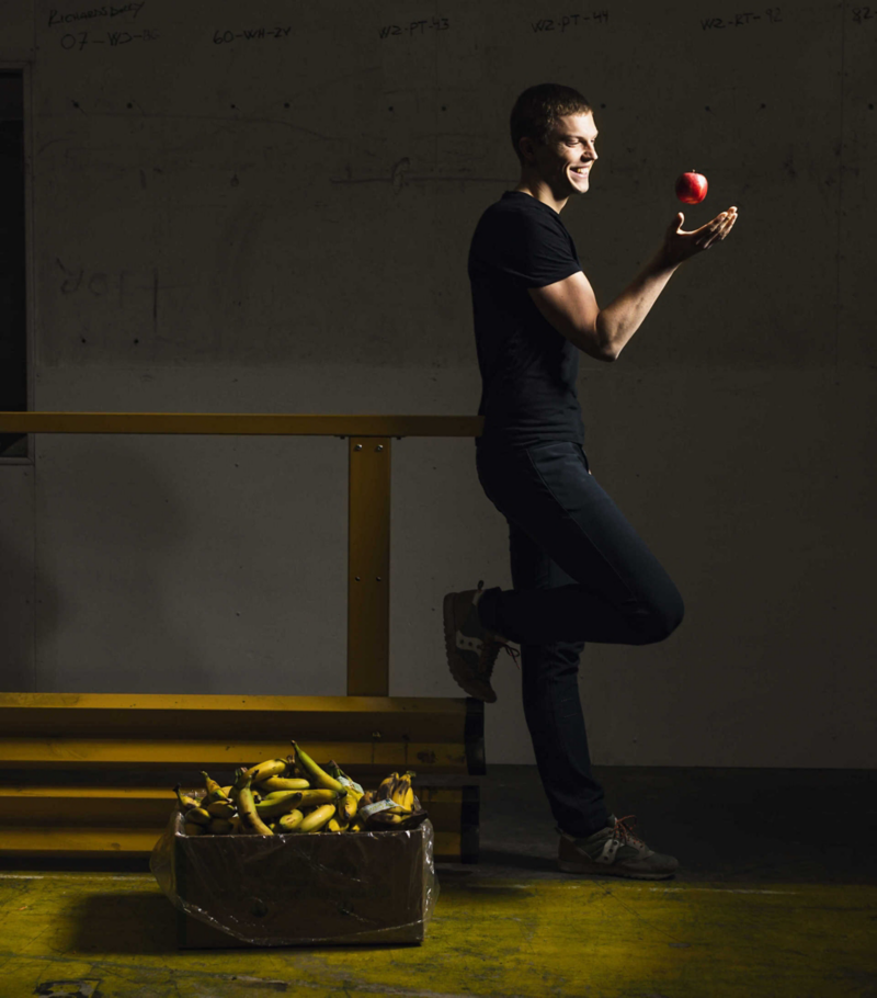 En person kastar ett äpple i luften, bredvid står en korg med bananer