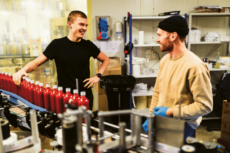 Två personer står vid en produktionslinje där flaskor fylls och hanteras