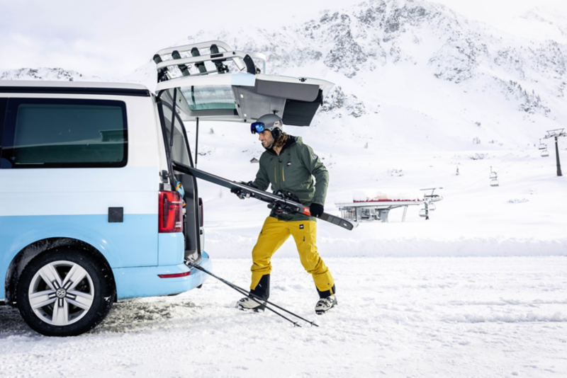  Un homme met des skis dans le coffre d'un van. Il est à la montagne, dans un paysage enneigé.