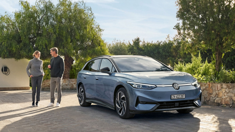 Volkswagen ID.7 vor einem Haus mit Wallbox geparkt, ein Paar spaziert daneben durch die Einfahrt im Grünen.