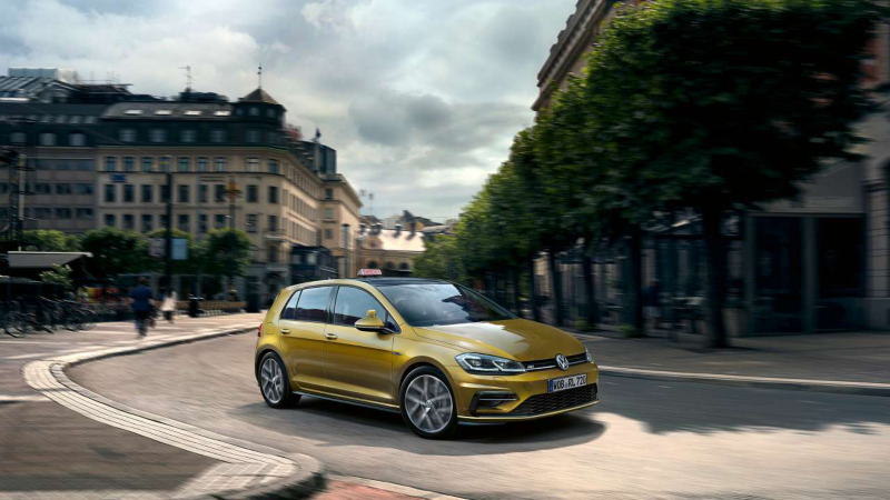 Une Volkswagen Golf de couleur or est garée au bord d'une rue urbaine bordée d'arbres et de bâtiments classiques, sous un ciel nuageux.