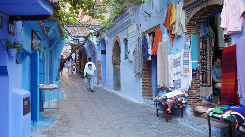 Ruelle étroite et peinte en bleu dans le souk de Chefchaouen, avec des textiles et des tapis suspendus et des personnes qui se promènent.