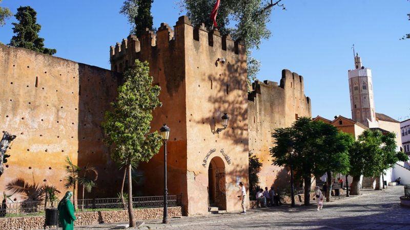 Fortification historique de la Kasbah au Maroc, en pierre ocre, avec des créneaux, une tour et des piétons sur la place.