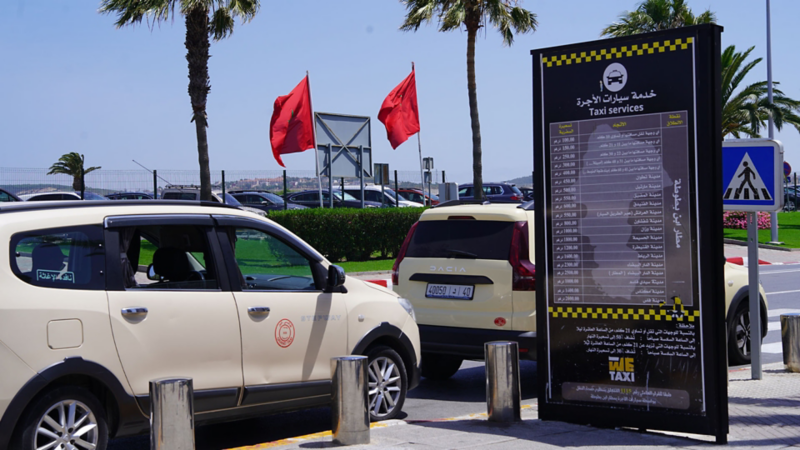 Taxis à l’aéroport au Maroc devant un grand tableau noir avec des prix écrits en arabe et des drapeaux marocains en arrière-plan.