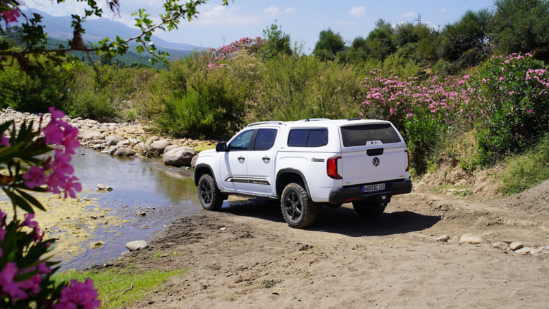 VW Amarok blanc, pick-up avec hardtop sur un chemin de terre à côté d’une rivière avec des arbustes d’oleandres roses en fleurs.