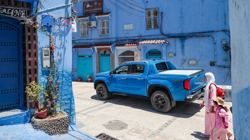 Un VW Amarok bleu est garé dans une ruelle étroite peinte en bleu à Chefchaouen, tandis qu’une mère et son enfant passent devant.