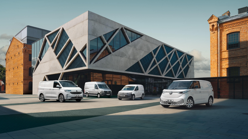 Four white VW vans are parked in front of a modern, angular building with triangular windows against a blue sky, conveying a sense of modernity and functionality