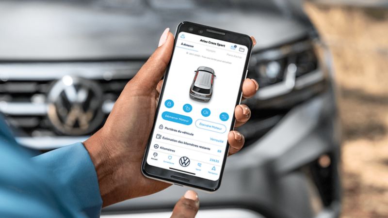 Une personne utilisant l'application Car-NetMD sur un téléphone devant un Atlas Cross Sport 2022 de Volkswagen argent pyrite métallisé.