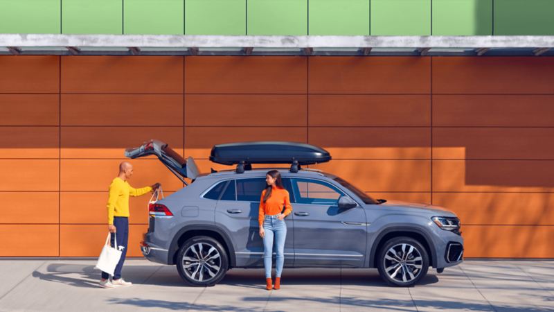 Une femme s’appuie sur un VWUS Atlas Cross Sport 2022 de Volkswagen gris pur tout en regardant un homme qui décharge le coffre.