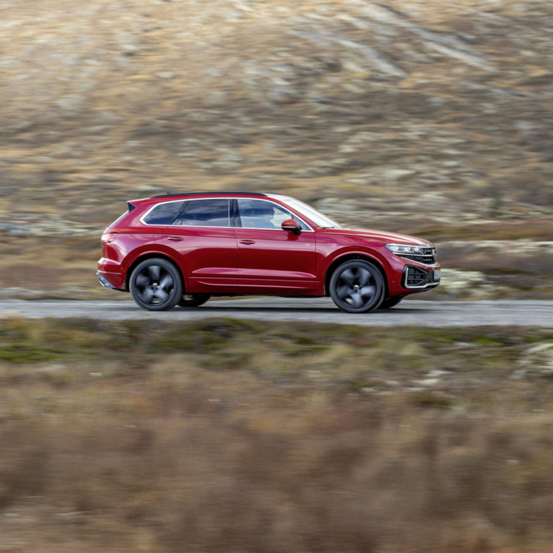 A VW Touareg Elegance parked in the sun, view from the side, behind it you can see the sea and the coast.