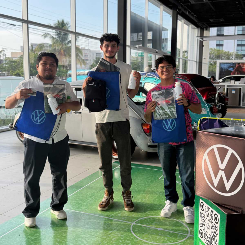 Tres hombres muestran artículos de Volkswagen frente a vochos de colección. 